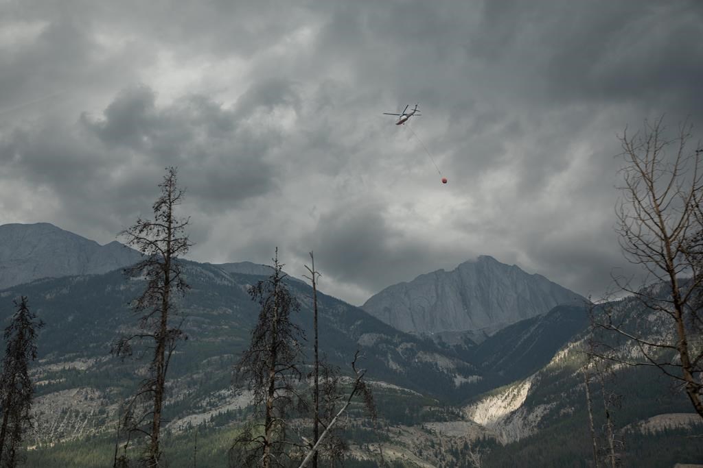 A helicopter buckets water onto smouldering fires outside of Jasper, Alta., on Friday July 26, 2024. Parks Canada says work to retrieve pets and restore various utility services continues around the Municipality of Jasper as an out of control wildfire burns nearby. THE CANADIAN PRESS/Amber Bracken