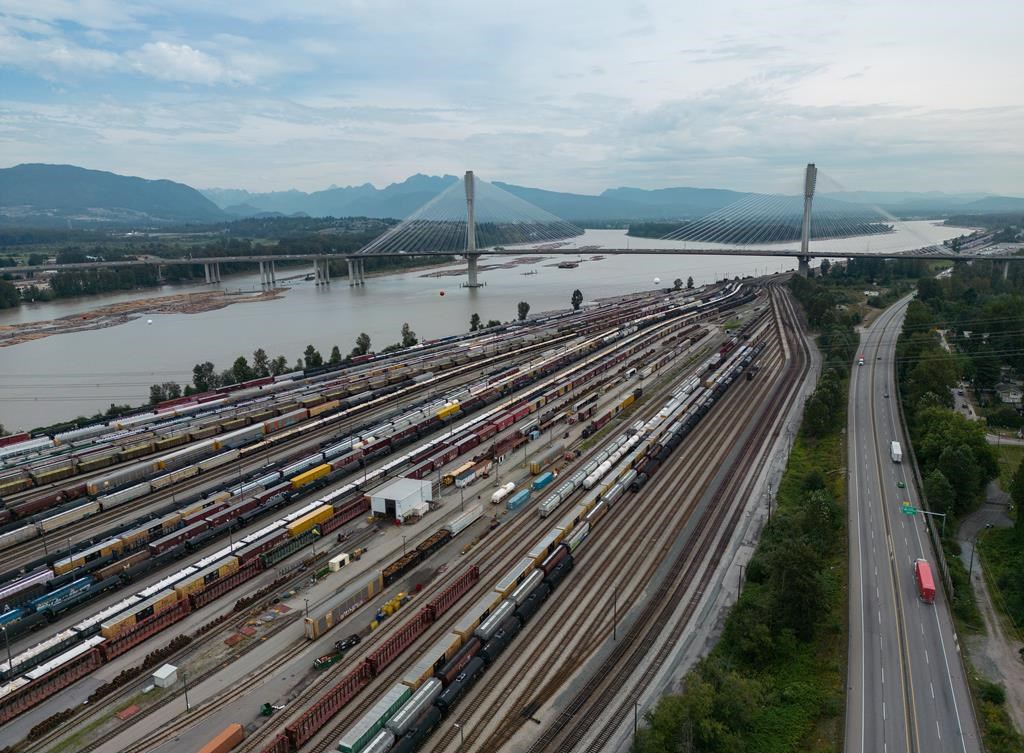 The ongoing labour dispute between Canada’s largest railway companies and their workers is driving up the cost of shipping by truck. Train cars are seen on the tracks in an aerial view at Canadian National Rail's Thornton Yard as trucks transport cargo containers on the highway, in Surrey, B.C., on Thursday, Aug. 22, 2024. THE CANADIAN PRESS/Darryl Dyck